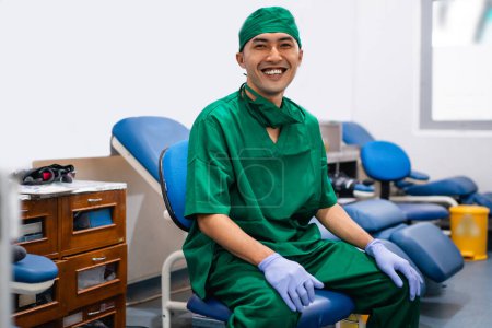Téléchargez les photos : Médecin masculin en uniforme vert et gants en caoutchouc sourit assis dans une salle de transfusion sanguine - en image libre de droit