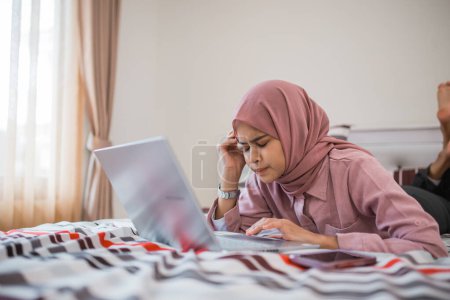 Photo for Woman in hijab gets dizzy while using laptop while lying on bed in room - Royalty Free Image
