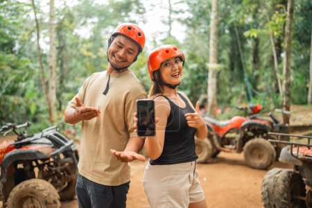 cute asian couple with savety equipment showing the blank phone on the camera with thumb up at atv tracking arena