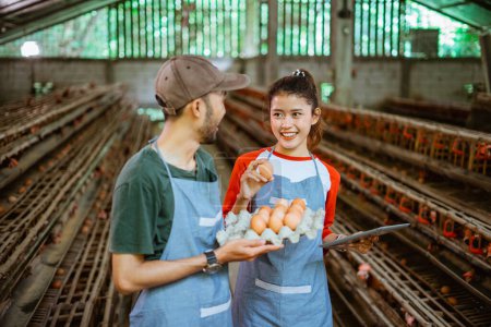 Téléchargez les photos : Travailleur femelle tenant un comprimé et prélevant des œufs sur un plateau en carton d'œufs transporté par un travailleur masculin dans une exploitation avicole - en image libre de droit