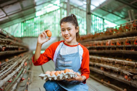 Téléchargez les photos : Belle agricultrice asiatique ramassant des œufs sur un plateau à la ferme de poulet - en image libre de droit