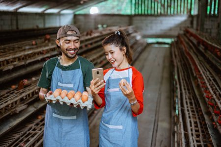 Téléchargez les photos : Sourire couple entrepreneur tenant plateau de carton d'oeuf et téléphone cellulaire stand à la ferme de poulet - en image libre de droit