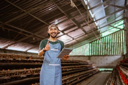 Téléchargez les photos : Sourire jeune entrepreneur asiatique à l'aide d'un tampon avec les pouces vers le haut sur la ferme de poulet - en image libre de droit