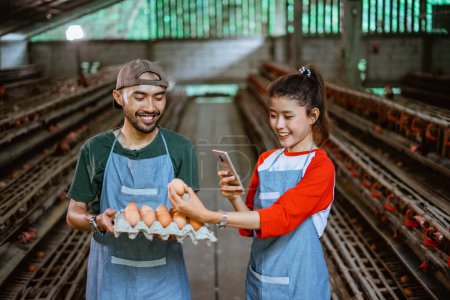 Téléchargez les photos : Couple d'entrepreneurs prendre des photos d'oeuf en utilisant un smartphone tout en tenant plateau de carton d'oeuf à la ferme de poulet - en image libre de droit