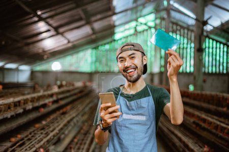 Téléchargez les photos : Homme d'affaires asiatique souriant montrant un livre de banque et tenant un téléphone debout dans une ferme de poulet - en image libre de droit