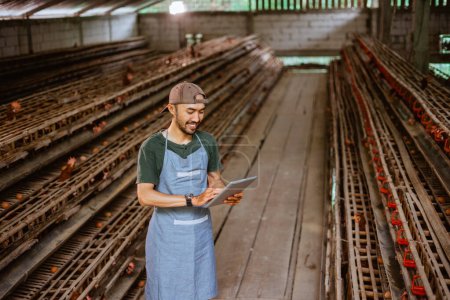 Téléchargez les photos : Jeune entrepreneur asiatique utilisant une tablette numérique debout dans la ferme de poulet avec de nombreuses poulaillers - en image libre de droit