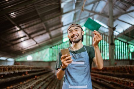 Téléchargez les photos : Entrepreneur montrant livre d'épargne et tenant smartphone debout à la ferme de poulet - en image libre de droit