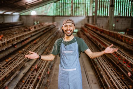 Téléchargez les photos : Jeune entrepreneur asiatique sourire avec geste de bienvenue debout dans la ferme de poulet avec de nombreuses poulaillers - en image libre de droit