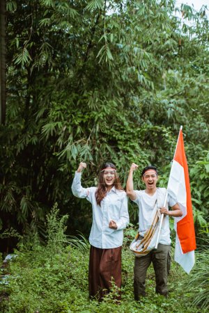 Téléchargez les photos : Un bel homme avec sarong saisir le drapeau indonésien à sa main debout à la forêt avec une belle femme en chemise blanche et serré leur bras - en image libre de droit