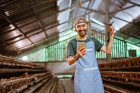 Photo for Excited young entrepreneur using a phone stands in chicken farm with many chicken coops - Royalty Free Image