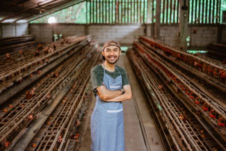 Téléchargez les photos : Jeune entrepreneur asiatique souriant avec les mains croisées se tiennent dans la ferme de poulet avec de nombreux poulaillers - en image libre de droit