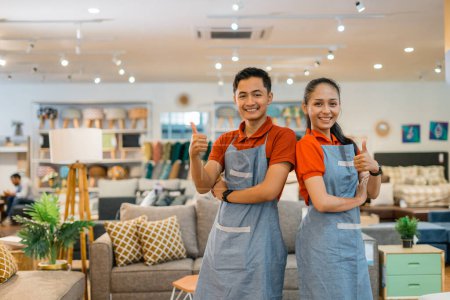 Téléchargez les photos : Hommes et femmes employés de magasin avec les pouces levés souriant à la caméra alors qu'ils se tenaient dans un magasin de meubles - en image libre de droit