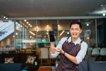 Téléchargez les photos : Assistant de magasin masculin dans le tablier avec geste de la main présentant téléphone portable devant le magasin de meubles - en image libre de droit