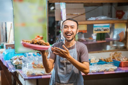 Téléchargez les photos : Jeune homme asiatique portant un plateau de collations avec un geste d'offrande dans un décor de stalle - en image libre de droit