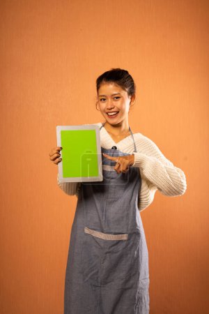 Téléchargez les photos : Asiatique fille portant tablier tenant tablette numérique avec doigt pointant vers écran vide sur fond isolé - en image libre de droit