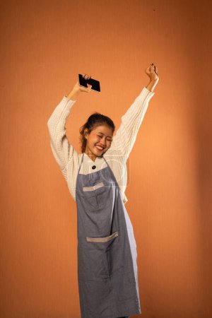 Téléchargez les photos : Excité fille asiatique portant tablier avec les mains en l'air et tenant téléphone cellulaire en arrière-plan isolé - en image libre de droit