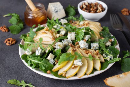 Salat mit Rucola, Birne, dor Blauschimmelkäse, Honig und Walnüssen im Teller auf grauem Tisch