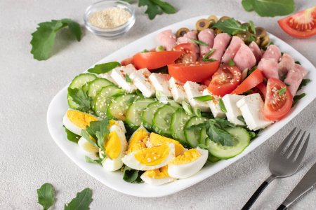 Healthy cobb salad with ham, feta cheese, cucumber, tomato, olives and eggs on white plate. Ketogenic lunch