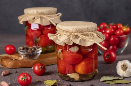 Pickled cherry tomatoes with garlic in two glass jars on brown background, stock for the winter