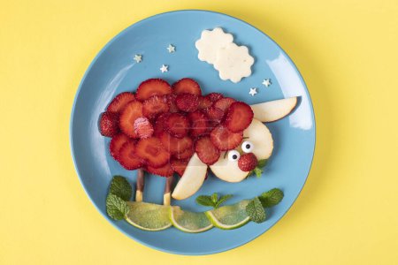 Fun food idea for kids - sheep animal with strawberry, apple and lime laid out on blue plate on yellow background, Top view