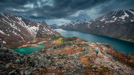 Foto de Woman hiking along turquoise and blue lakes in mountain landscape on hike of Knutshoe in Jotunheimen National Park in Norway, mountains of Besseggen in background, dramatic cloudy sky - Imagen libre de derechos