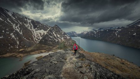 Foto de Woman hiking along blue lake in mountain landscape of Knutshoe in Jotunheimen National Park in Norway, mountains of Besseggen in background, dramatic cloudy sky - Imagen libre de derechos