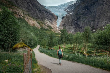 Téléchargez les photos : Woman hiking along path to Briksdalsbreen glacier in the mountains of Jostedalsbreen national park in Norway, green meadows with flowers in foreground - en image libre de droit