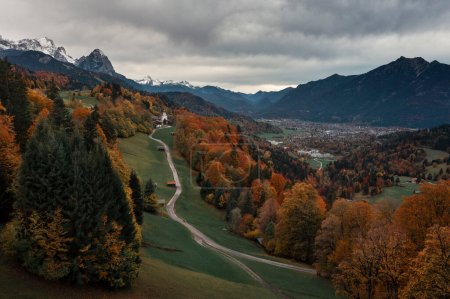 Photo for Bavarian Alps with church of Wamberg in Garmisch-Partenkirchen during autumn, snow-covered mountains in the background, dramatic cloudy sky, colored leaves and trees and huts and road in foreground, Bavaria Germany - Royalty Free Image