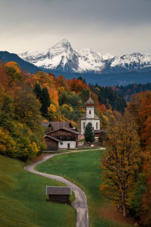 Photo for Bavarian Alps with church of Wamberg in Garmisch-Partenkirchen during autumn, snow-covered mountains in the background, dramatic cloudy sky, colored leaves and trees and huts and road in foreground, Bavaria Germany - Royalty Free Image