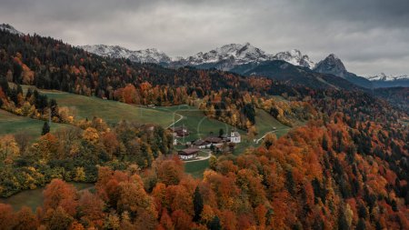 Photo for Bavarian Alps with church of Wamberg in Garmisch-Partenkirchen during autumn from above, snow-covered mountains in the background, dramatic cloudy sky, colored leaves and trees and road in foreground, Bavaria Germany - Royalty Free Image