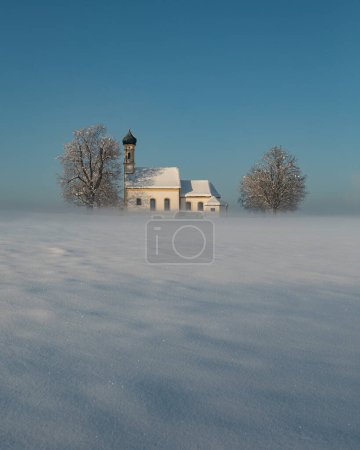 Foto de Bavarian church of Raisting with trees and snow and mist during winter, snow field in the foreground, blue sky day, Bavaria Germany - Imagen libre de derechos