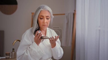 Caucasian old senior mature woman lady 60s grandmother model in bathrobe in bath touching gray long smooth thin hair worry about lost hairline weak baldness aging problem alopecia hairstyle sickness