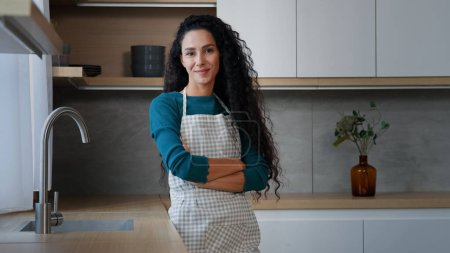 Photo for Smiling young woman maid housewife mistress baker housekeeper with long curly hair wearing checkered apron standing with folded arms confident chef posing in modern domestic kitchen cuisine interior - Royalty Free Image
