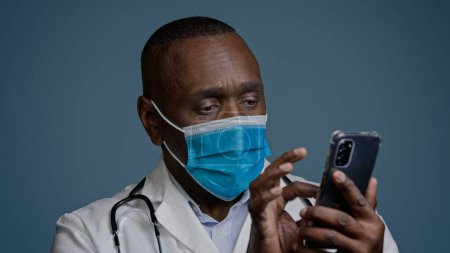 Photo for Close-up pensive thoughtful african american man doctor in protective mask holds phone studies new information on Internet ponders uses medical mobile application remotely consults patient online - Royalty Free Image