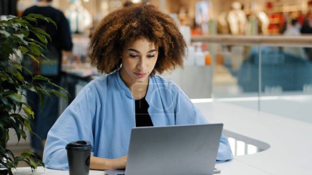 Photo for Front view focused millennial african american girl business woman freelancer manager specialist sitting at table in cafe working with laptop answering message writes report study drinking coffee - Royalty Free Image