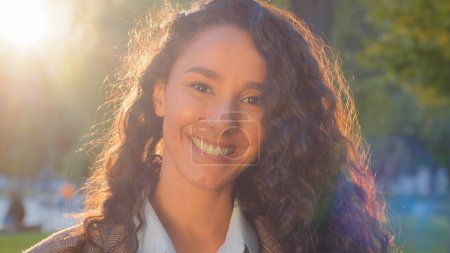 Photo for Beautiful female lady girl looks at camera with toothy smiledental laugh posing in sun lights outdoors. Woman with long black curly hair in sunbeams in park. Attractive model with flying curls . High - Royalty Free Image