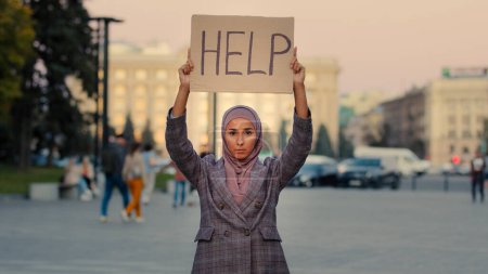 Téléchargez les photos : Triste solitaire pauvre fille musulmane porte hijab femme se tient en ville en regardant la caméra montre bannière en carton texte aide a besoin de travail de protection, concept de problème de discrimination de chômage. Haut - en image libre de droit