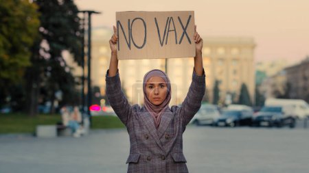 Téléchargez les photos : Jeune femme musulmane sérieuse fille islamique dans le hijab militant féminin se tient dans la rue de la ville protestant à l'extérieur tenant bannière signe avec texte pas de vax contre la vaccination interdiction de refuser arrêter la vaccination protestation - en image libre de droit