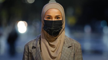 Foto de Mujer islámica de cerca con máscara médica posando en la ciudad nocturna. Musulmana mujer de negocios con hijab enmascarado chica de pie mirando a la cámara de noche fondo urbano al aire libre pandemia coronavirus covid - Imagen libre de derechos