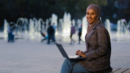 Téléchargez les photos : Occupé fille étudiante musulmane réussie dans hijab jeune femme d'affaires islamique en utilisant l'application pour ordinateur portable travaillant dans les fontaines de la ville arrière-plan en regardant la caméra sourire heureux. Femme ethnique avec ordinateur sans fil - en image libre de droit