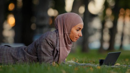 Téléchargez les photos : Amicale musulmane femme d'affaires islamique fille musulmane dans le hijab gazon couché pelouse dans le parc en plein air enseignante mentor en ligne faire une conférence de chat vidéo parler webcam en utilisant un ordinateur portable communication à distance - en image libre de droit