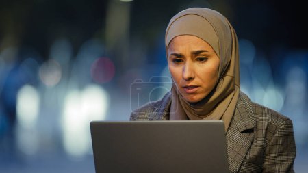 Foto de Retrato de musulmana mujer de negocios usuario en hijab se sienta en la ciudad de la noche al aire libre trabajando con el ordenador portátil preocuparse por el problema de mirar victorias de la computadora dice que sí. Estudiante islámica chica consigue oferta en línea ganar - Imagen libre de derechos