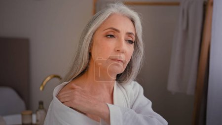 Caucasian granny grandmother old model female elderly 60s Caucasian lady woman in bathrobe in bathroom smears shoulders back and neck chest with lotion body cream oil moisturizing soft skin body care