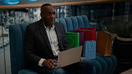Photo pour Afro-Américain homme d'affaires travailleur entrepreneur client homme dans le centre commercial assis sur le canapé avec des sacs achats à l'aide d'un ordinateur portable acheter boutique en ligne choisir des marchandises acheter commande e-commerce - image libre de droit