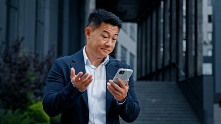 Photo for Sad Asian businessman in city mobile problem with telephone. Middle-aged CEO employer entrepreneur outdoor browsing phone feeling upset with broken mistake error online cellular bad connection lost - Royalty Free Image