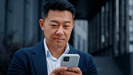 Photo for Close up Asian 40s businessman with smartphone texting browsing smiling looking at camera standing outdoors in city. Middle-aged man CEO has business chat using mobile phone check news in social net - Royalty Free Image