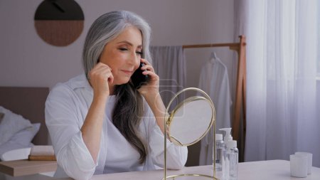 Foto de Senior de pelo gris de edad de 60 años señora hablando de teléfono móvil conversación mirando espejo reflexión comprar crema facial por teléfono inteligente disfrutar arrugado hermoso envejecimiento perfecta piel lisa - Imagen libre de derechos