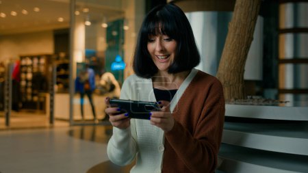 Woman lady female gamer girl indoors in shopping mall sitting playing with mobile phone in video online game in net smartphone gaming app emotional enjoy play competition win celebrate victory winning