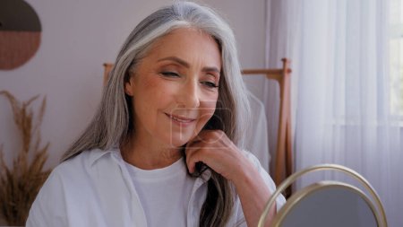 Photo for 50s yeas old lady with gray hair enjoying skin moisture looking at mirror reflection touch face cream applying 60s age senior mature woman check cosmetology results facial cosmetics touching cheeks - Royalty Free Image