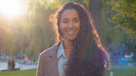 Photo for Female portrait outdoors beautiful smiling cheerful carefree young woman brunette girl lady with long curly hair and white teeth stands in sunshine sunbeams sun in park looking into camera friendly - Royalty Free Image